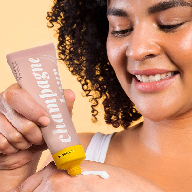 Person applying champagne-colored lotion to their shoulder with a beige background