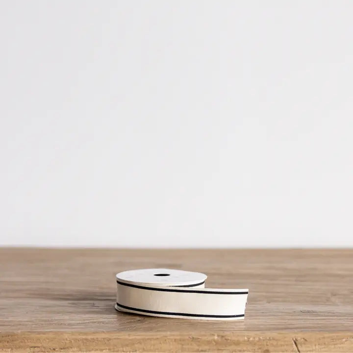 Cream woven ribbon spool with narrow black edge trim displayed on a wood tabletop.