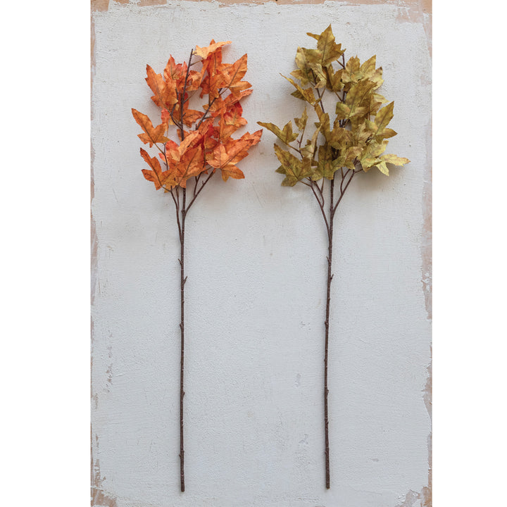 Two artificial leaf branches, one orange and one gold, on a textured white background.