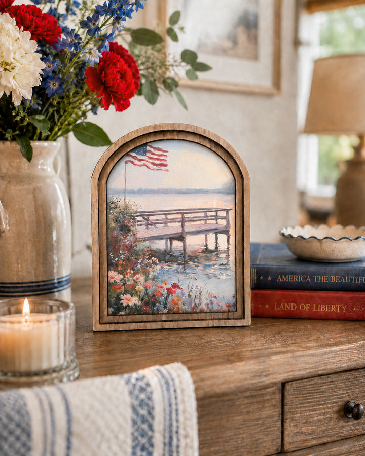Decorative scene with a framed picture of a dock and flowers, a vase of flowers, and books on a wooden surface.