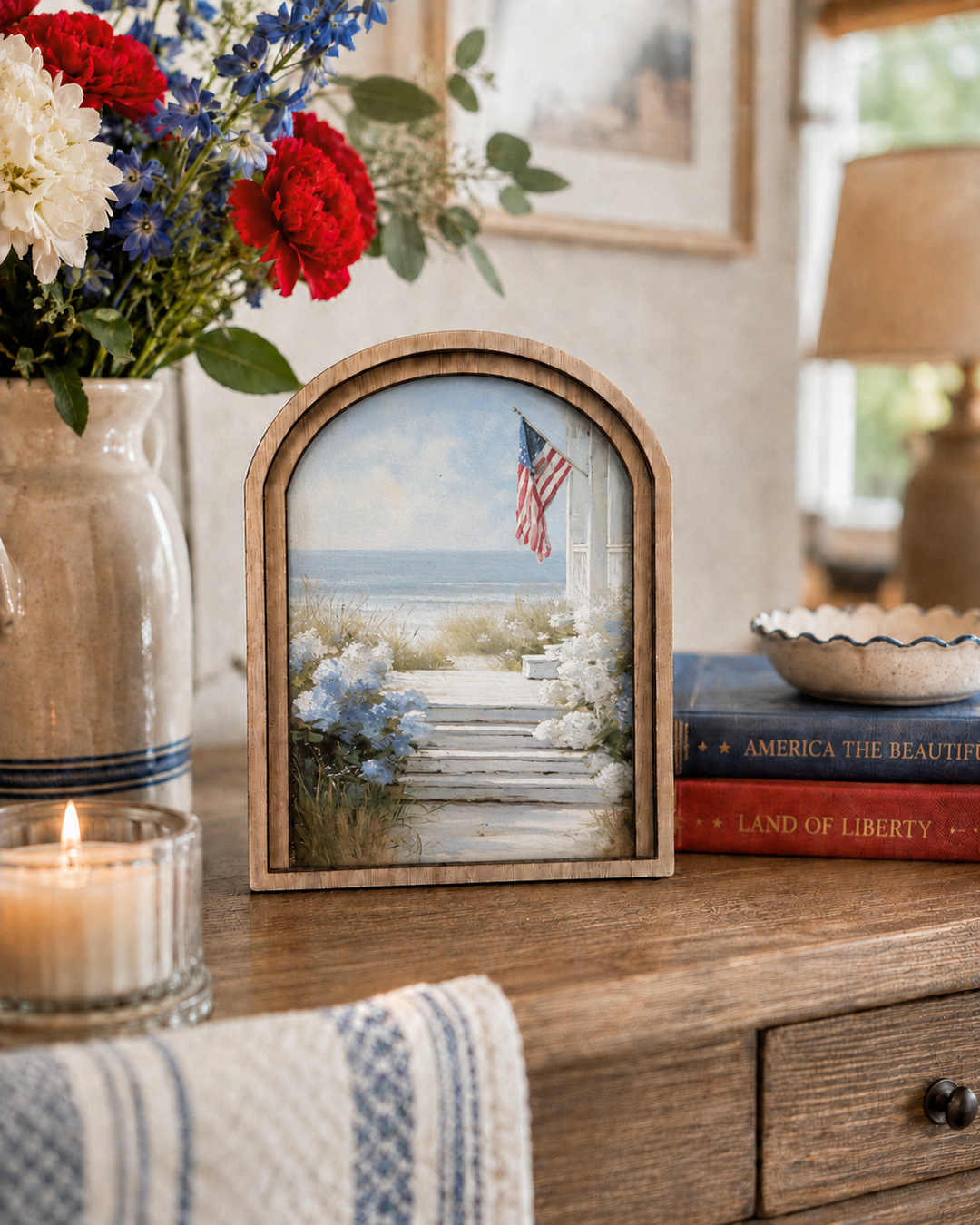 Decorative scene with a framed picture of a beach, American flag, flowers, and books on a wooden surface.