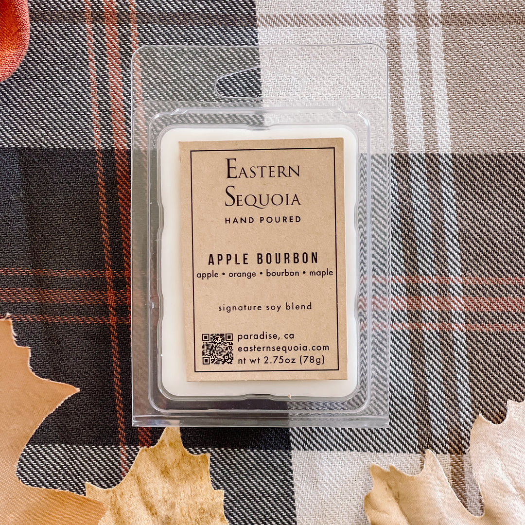 A wax melt pack labeled 'Eastern Sequoia' with 'Apple Bourbon' scent, placed on a plaid fabric background with leaves around it.