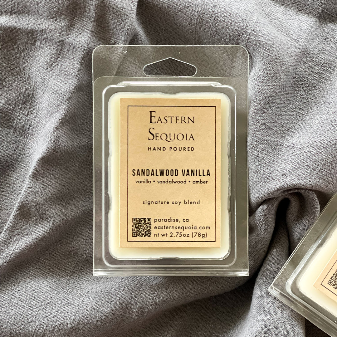 A clear plastic package containing Sandalwood Vanilla wax melts with a beige label that reads 'Eastern Sequoia - Hand Poured - Sandalwood Vanilla - Signature Soy Blend'. The package is placed on a grey fabric surface.