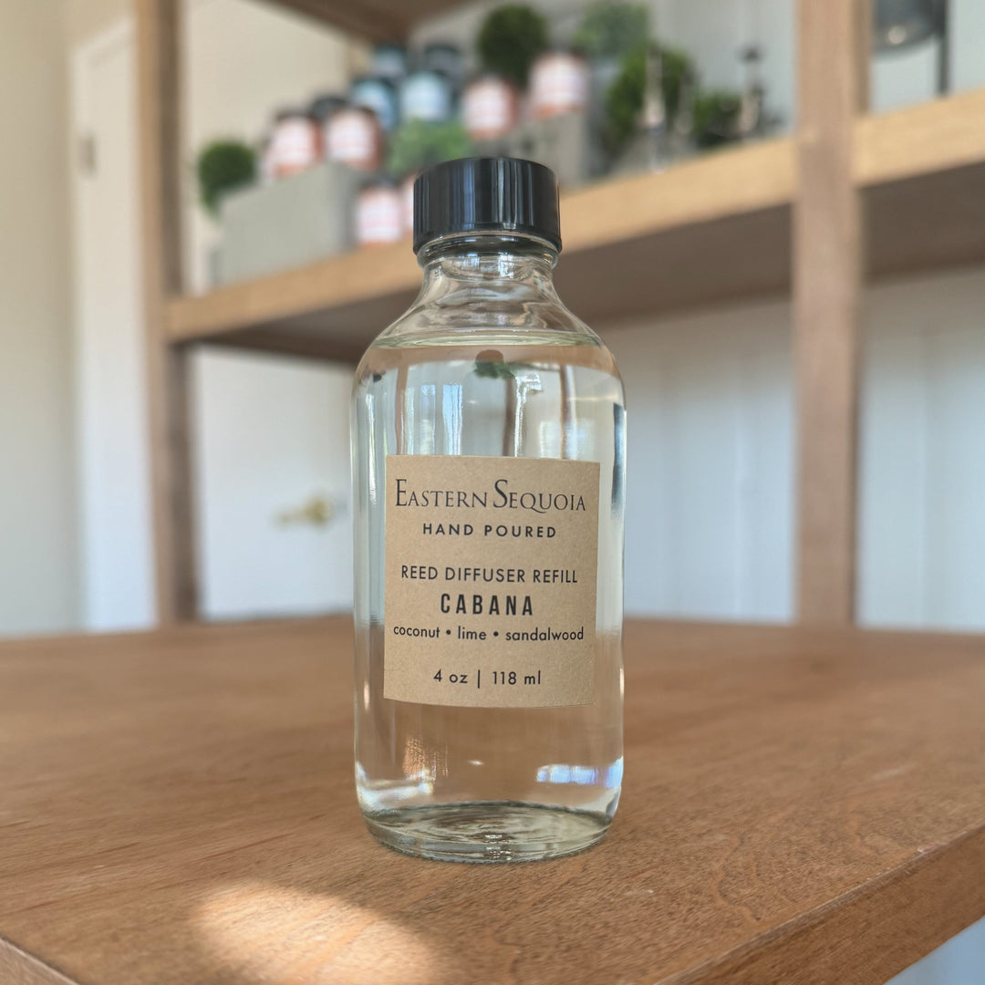A clear glass bottle of Cabana reed diffuser refill on a wooden surface with blurred shelves in the background.
