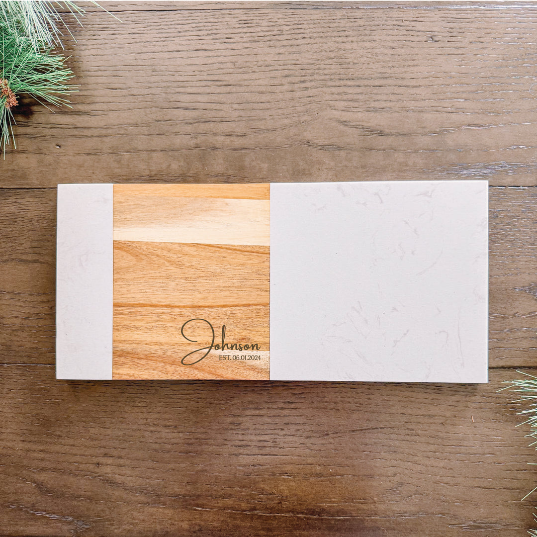 A personalized charcuterie board with a marble and acacia wood surface, featuring a engraved family name.