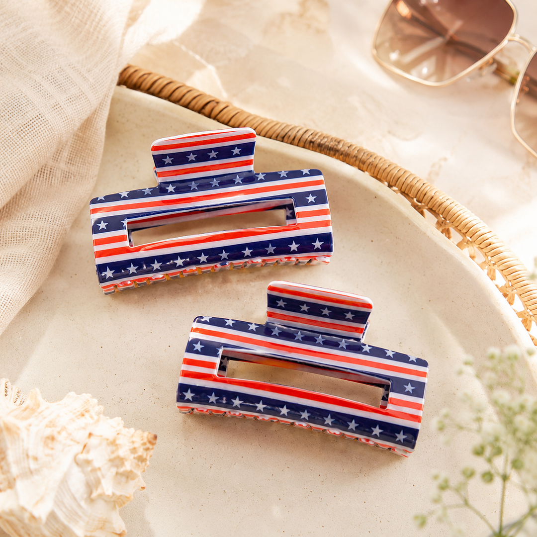 Two American flag patterned hair clips on a textured surface with a shell and sunglasses in the background.
