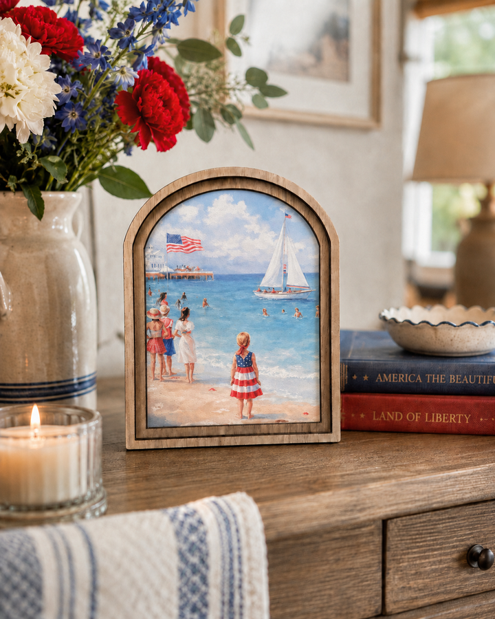 Small framed painting of a beach scene with people and an American flag, placed on a wooden surface with flowers and books.