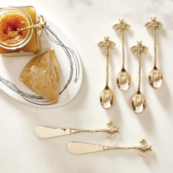 Set of gold spoons and butter knives with decorative handles on a white surface, next to a plate with a pastry and jam.