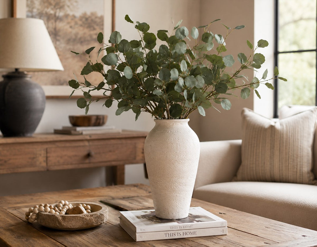 Textured white ceramic farmhouse vase with eucalyptus branches styled on a rustic wood coffee table in a warm neutral living room.