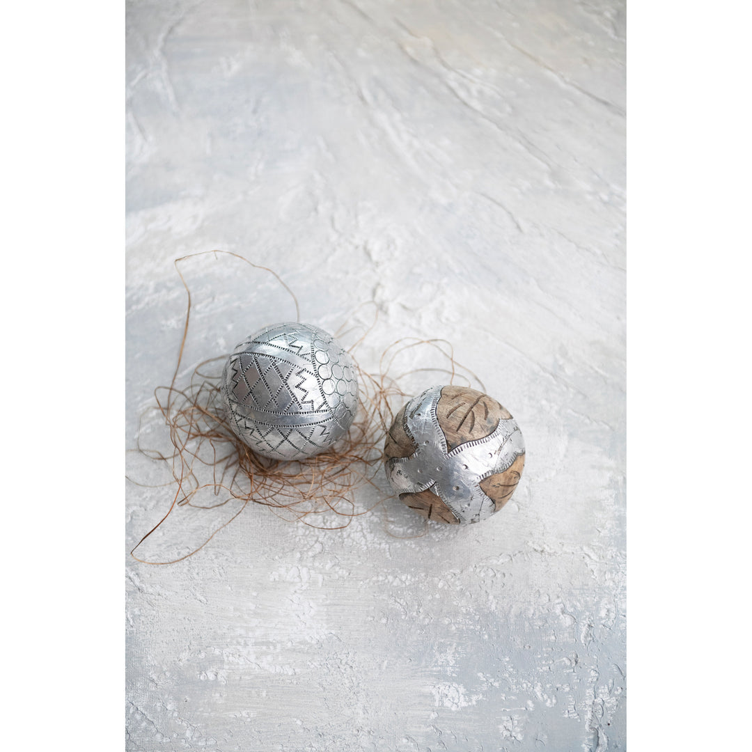 Two decorative spherical ornaments with a rustic, hand-painted look. One ornament is metallic silver with a geometric pattern, resting on a bed of brown twine.