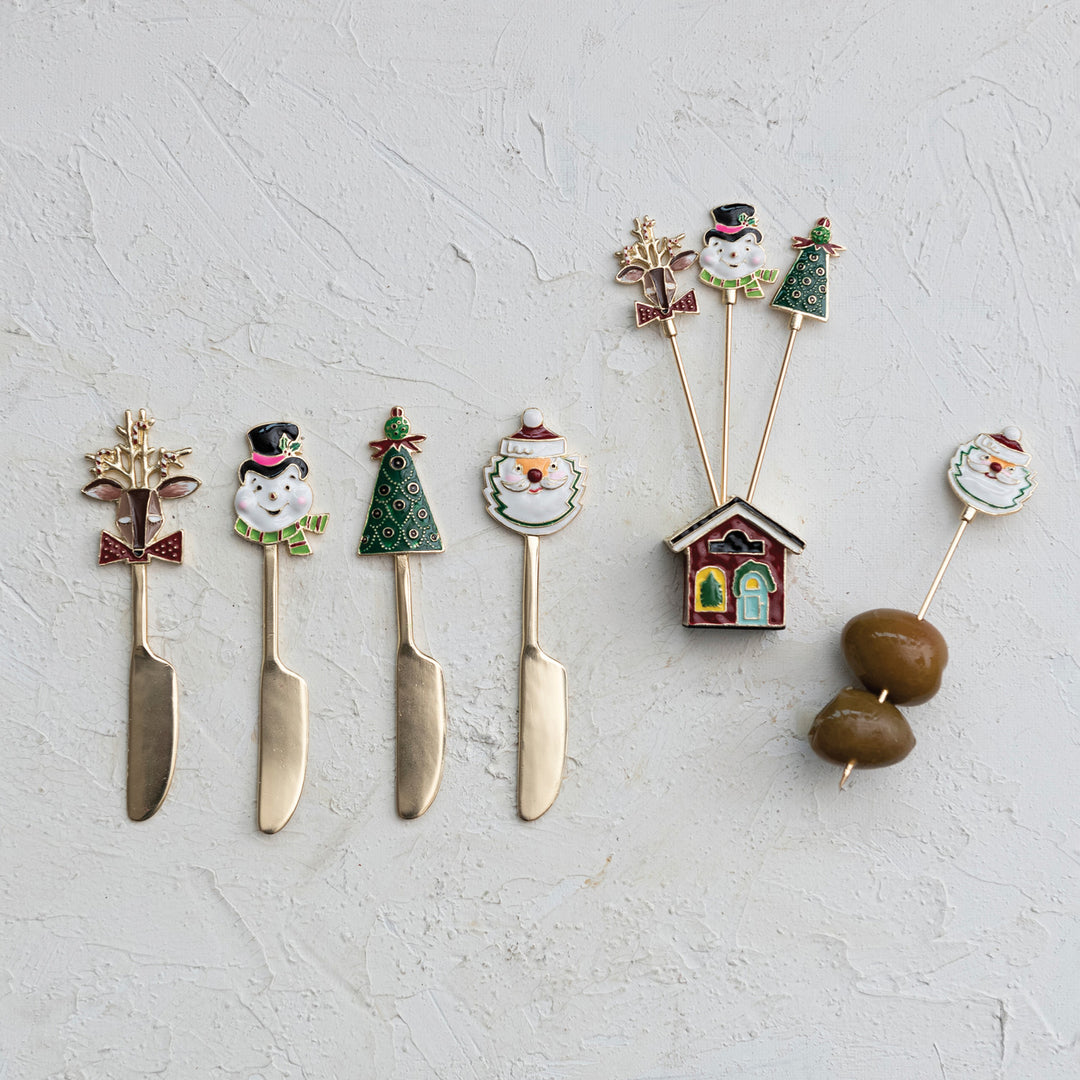 A set of decorative cheese knives with holiday-themed enameled handles, displayed on a textured light gray background. The handles feature designs including a snowflake, a snowman, a Christmas tree, a Santa face, a small house, and two brown olives.