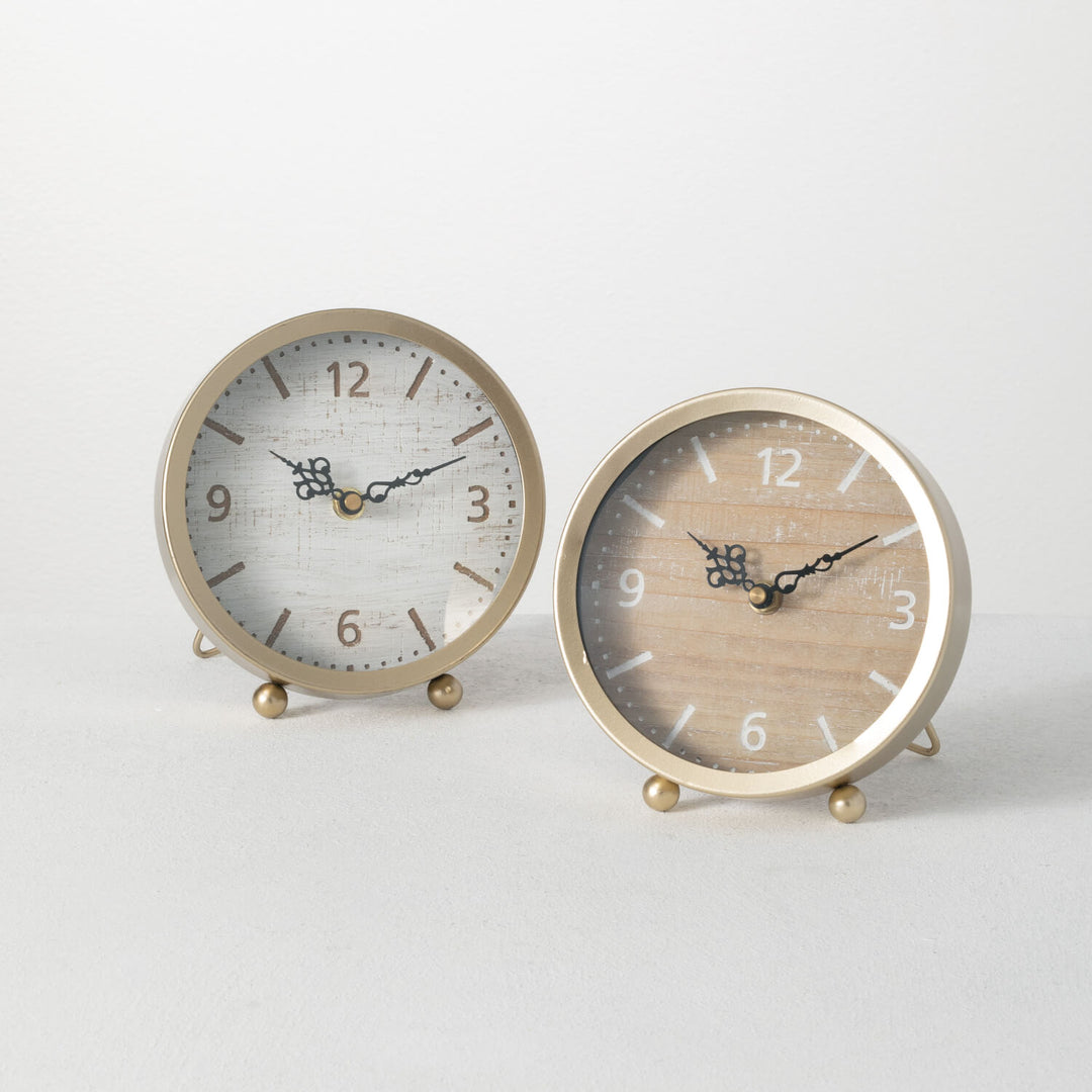Two round desk clocks with a wooden appearance and black hands on a white background, one with a gold frame, the other with a silver frame.
