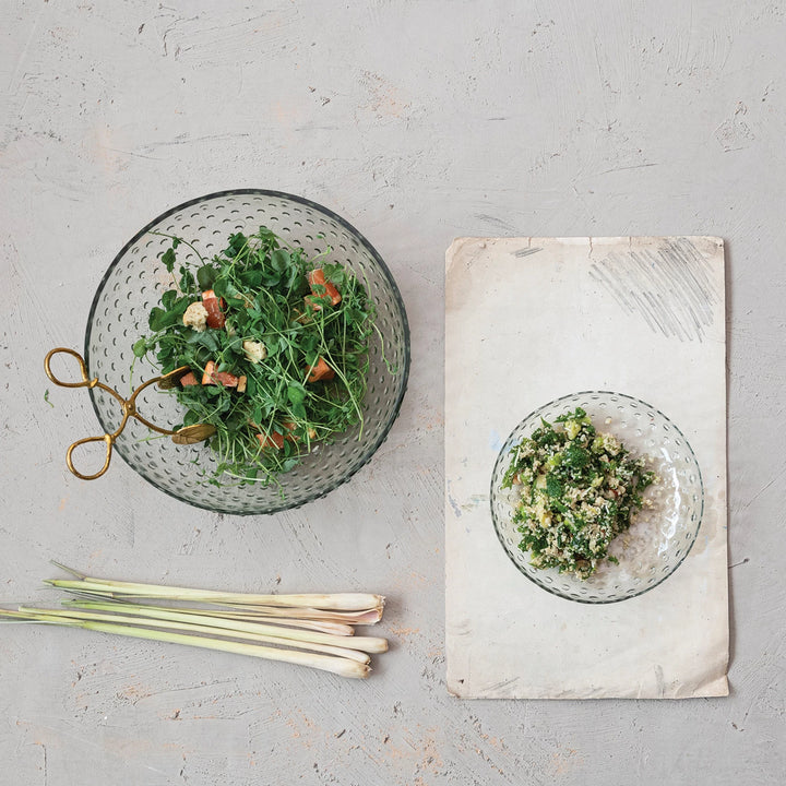 A clear glass serving bowl with a hobnail design, alongside a smaller recycled glass bowl, lemongrass stalks, and a cloth on a grey background.