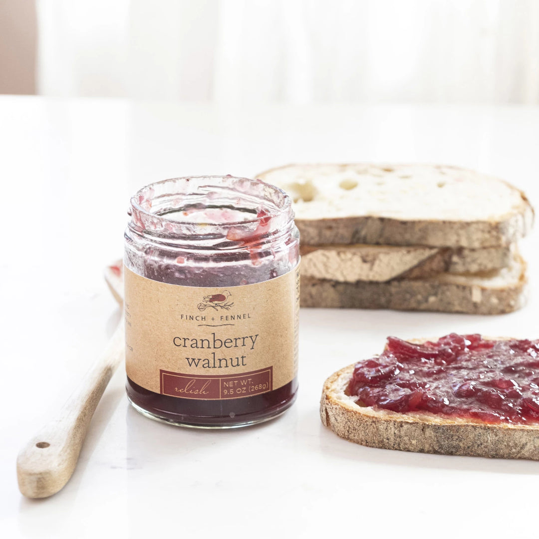 A jar of Finch + Fennel Cranberry Walnut Relish with a piece of bread with cranberry spread on it, placed on a white surface.