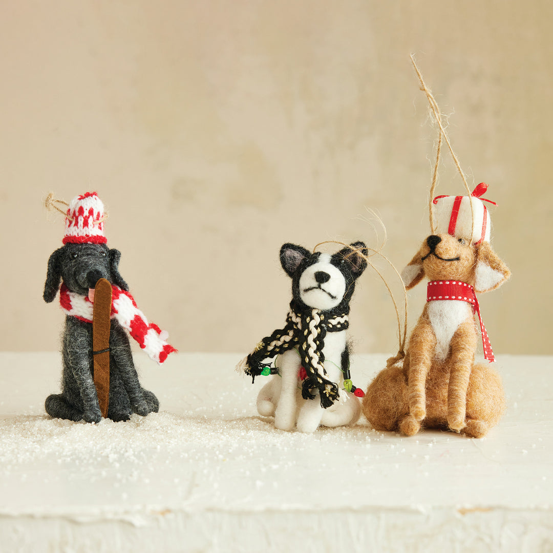 Three small felt dog ornaments dressed in holiday attire, including a black dog in a red-and-white hat and scarf, a black-and-white dog in a black-and-white scarf, and a tan dog in a red-and-white hat and scarf, arranged on a snowy surface against a soft beige background.