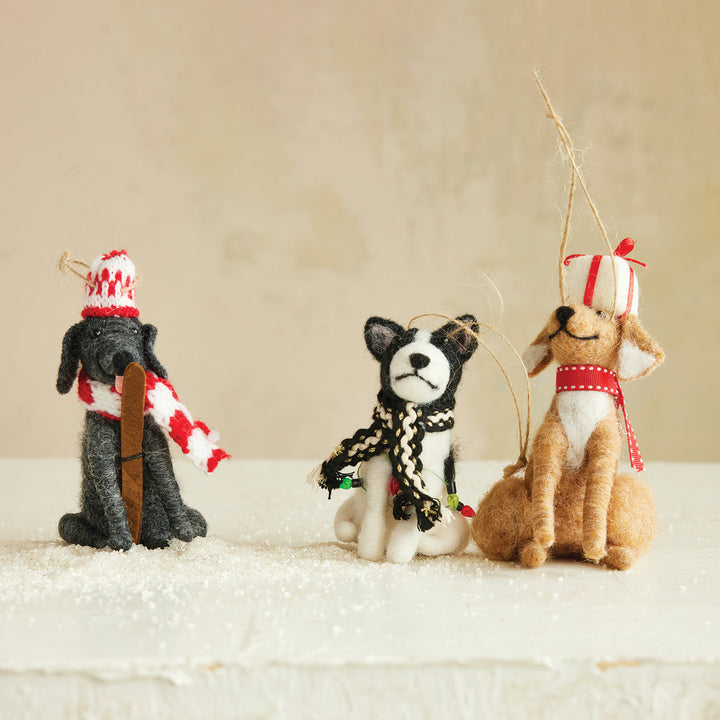 Three small felt dog ornaments dressed in holiday attire, including a black dog in a red-and-white hat and scarf, a black-and-white dog in a black-and-white scarf, and a tan dog in a red-and-white hat and scarf, arranged on a snowy surface against a soft beige background.