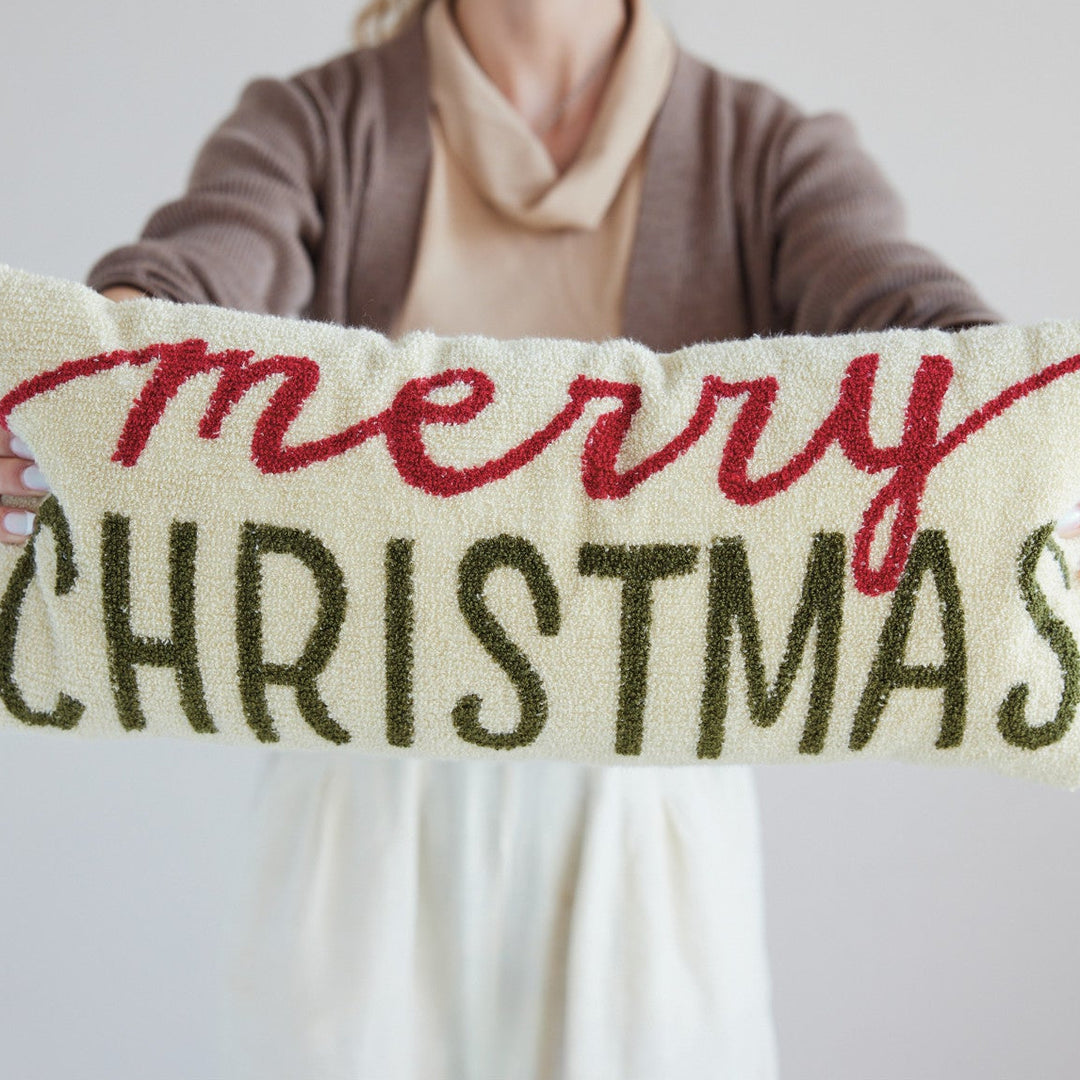 A person holds a long rectangular decorative pillow with the words “merry CHRISTMAS” in red and dark green embroidery on a cream background, against a plain light gray backdrop.