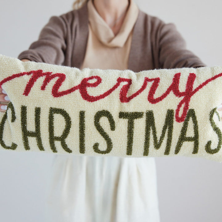 A person holds a long rectangular decorative pillow with the words “merry CHRISTMAS” in red and dark green embroidery on a cream background, against a plain light gray backdrop.