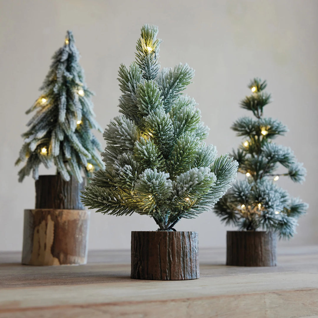 Three small artificial flocked Christmas trees with warm white string lights, displayed on rustic wood log bases against a soft beige background.